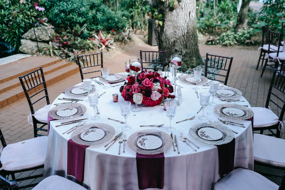 Reception table with rich floral centerpiece, elegant place settings, and deep-toned accents, styled in a garden setting for a sophisticated destination wedding in Florida.

