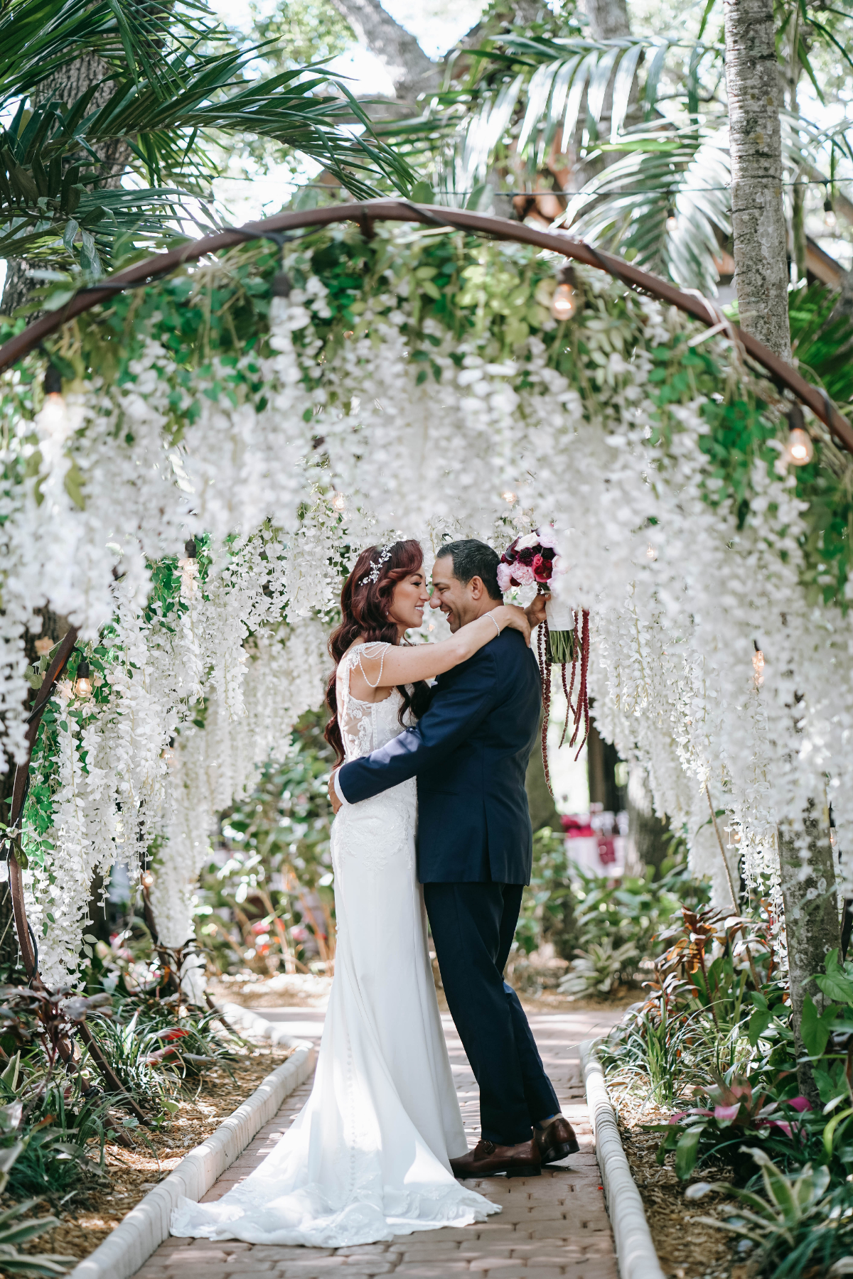 Couple embracing beneath a floral arch with cascading white blooms in a tropical garden pathway, creating a romantic ceremony setting for a destination wedding in Florida.