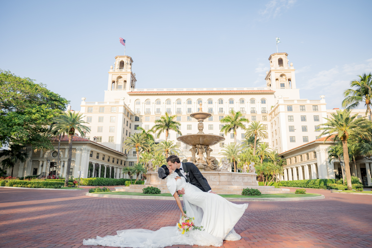 Bride and groom sharing a kiss in front of a grand fountain and palm-lined resort entrance, highlighting the iconic luxury and scale of a destination wedding in Florida.