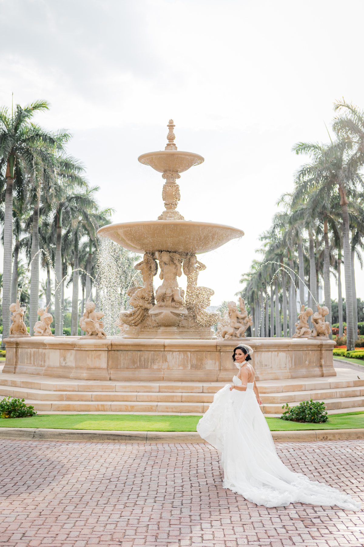 Bride walking gracefully in front of a large fountain framed by palm trees, showcasing a sunlit, luxurious outdoor setting ideal for a destination wedding in Florida.