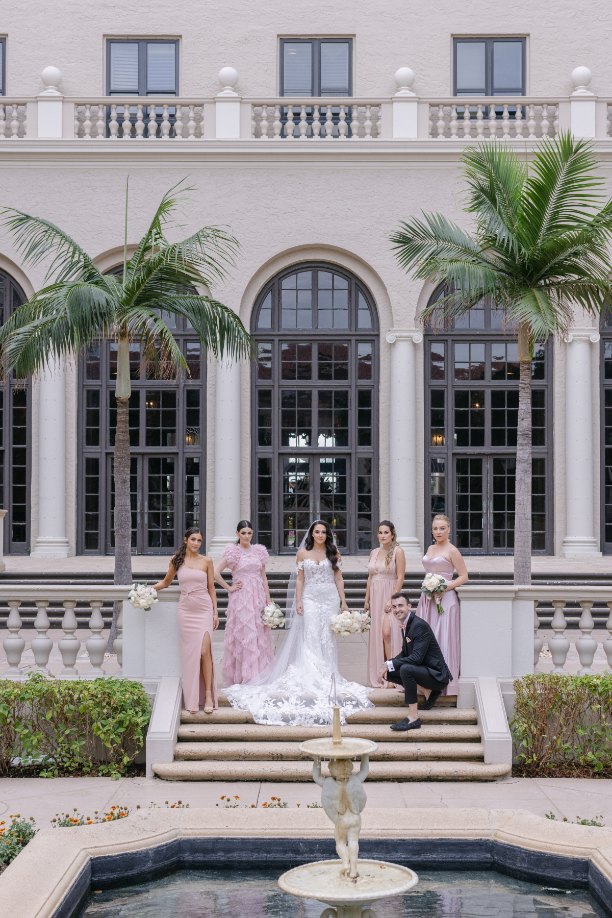 Wedding party posing on steps in front of a Mediterranean-style venue with arched windows and palm trees, reflecting the stylish architecture of a destination wedding in Florida.