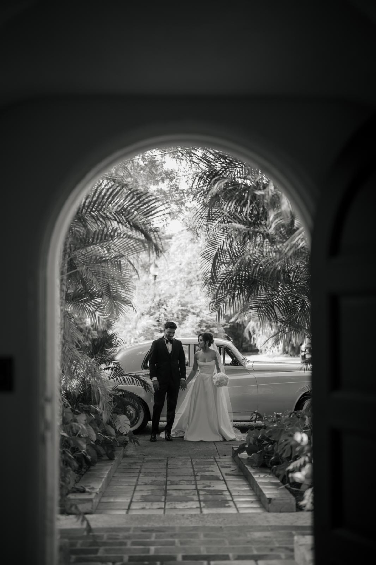 Black and white portrait of a couple standing by a vintage car framed by an arched doorway and tropical foliage, adding a timeless, editorial feel to a destination wedding in Florida.