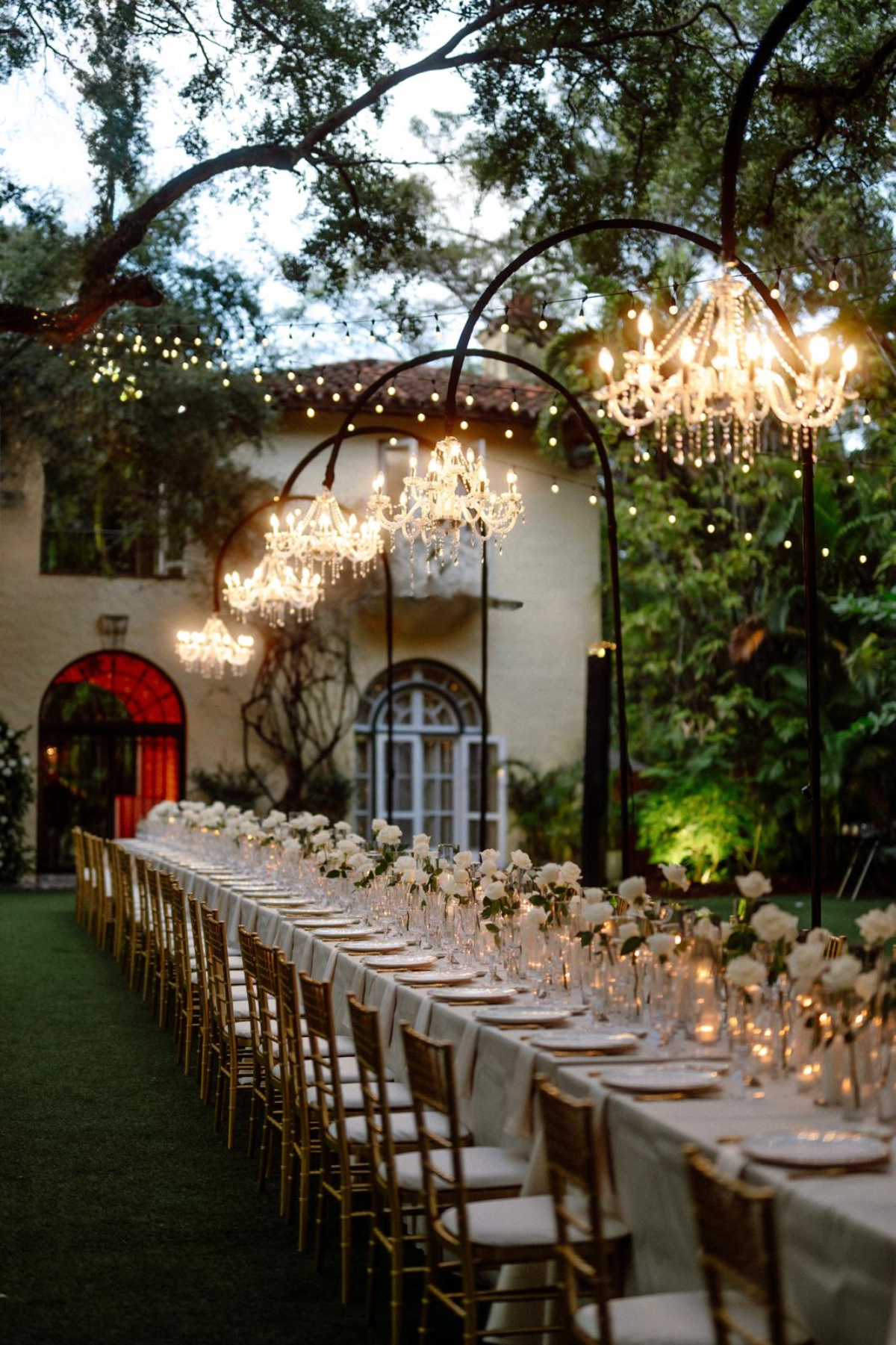 Evening reception setup at a villa courtyard with glowing chandeliers, bistro lights, and long banquet tables, evoking a warm, romantic ambiance for a destination wedding in Florida.