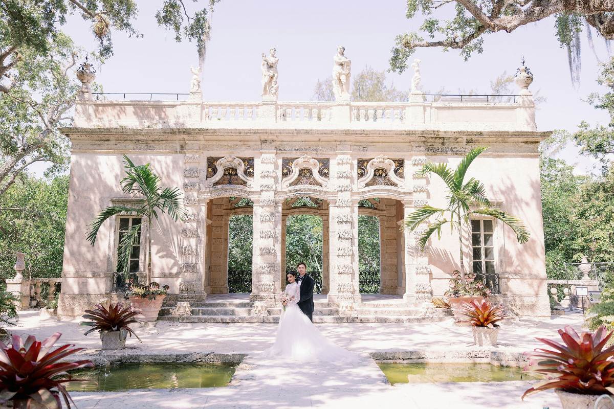 Newlywed couple posing beneath an ornate stone pavilion surrounded by lush greenery and water features, showcasing the architectural beauty and romantic atmosphere of a destination wedding in Florida.