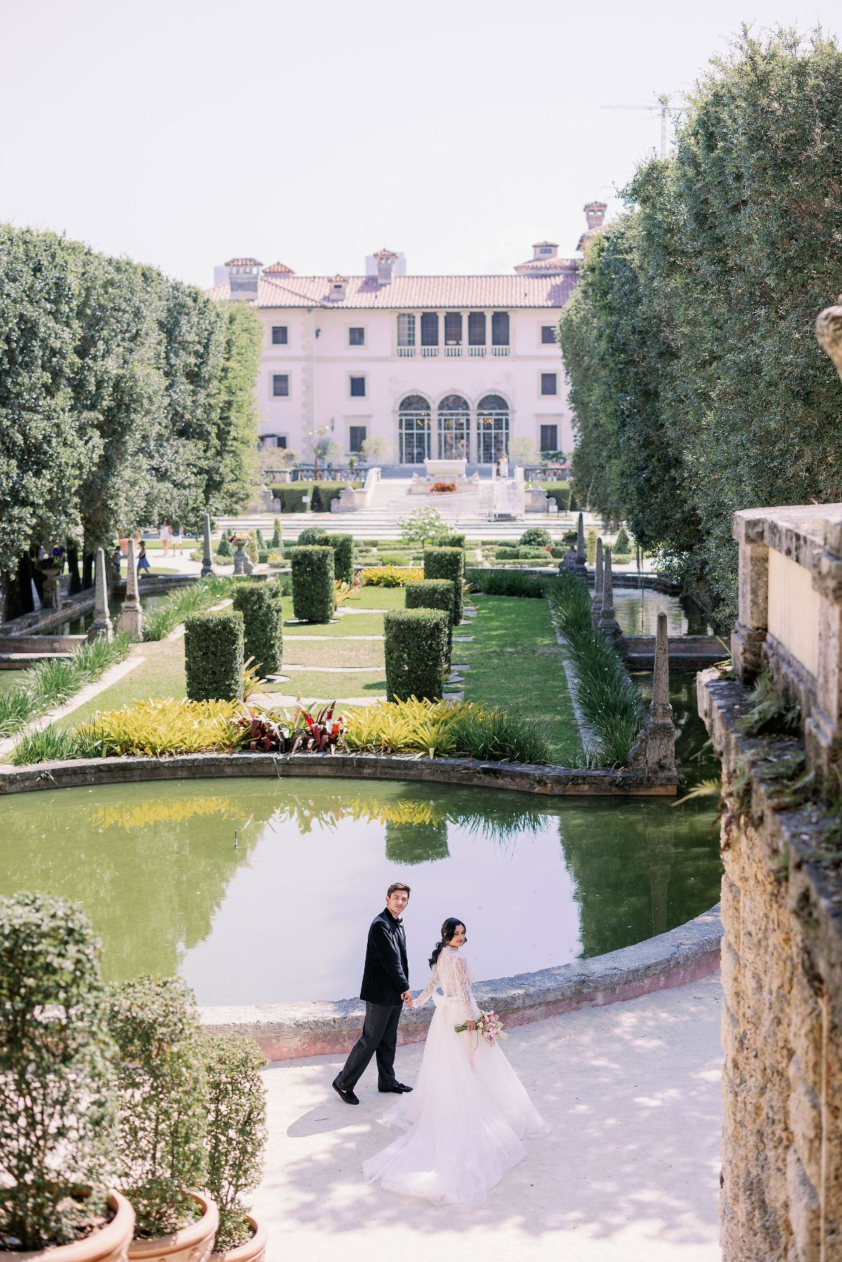 Bride and groom walking hand in hand beside a reflecting pool at a grand historic estate with manicured gardens, capturing the elegance and European charm of a destination wedding in Florida venue.